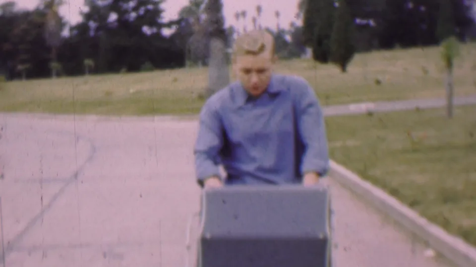 A young man in a blue shirt appears to concentrate on pushing a dark box or container along a paved park path in the 1955 San Diego home-movie scene.