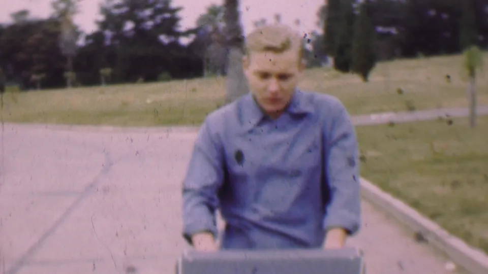 The final frame shows a closer view of the young man in a blue shirt with his hands near a dark box or container as he moves along the paved path in the park.
