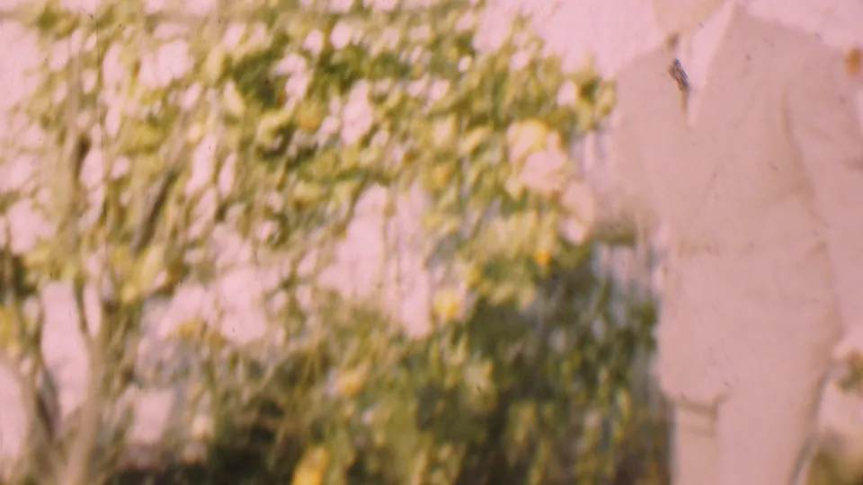 A cropped figure in a suit appears at the right edge of a bright garden scene with leafy shrubs and a pale pink wall, consistent with the San Diego 1955 clip metadata.