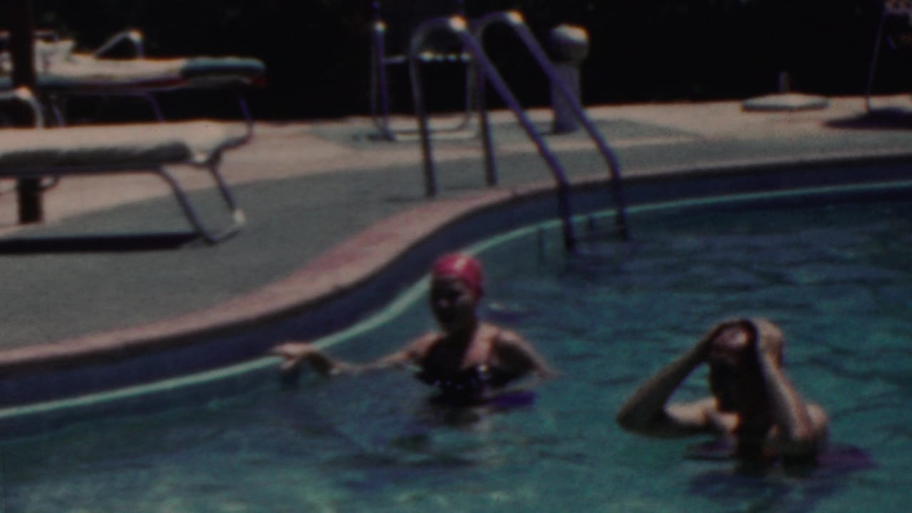Two adults swim near the edge of a pool in the 1949 Seaside, New Jersey home-movie footage, with a red swim cap, lounge chairs, and a metal ladder visible on the deck.