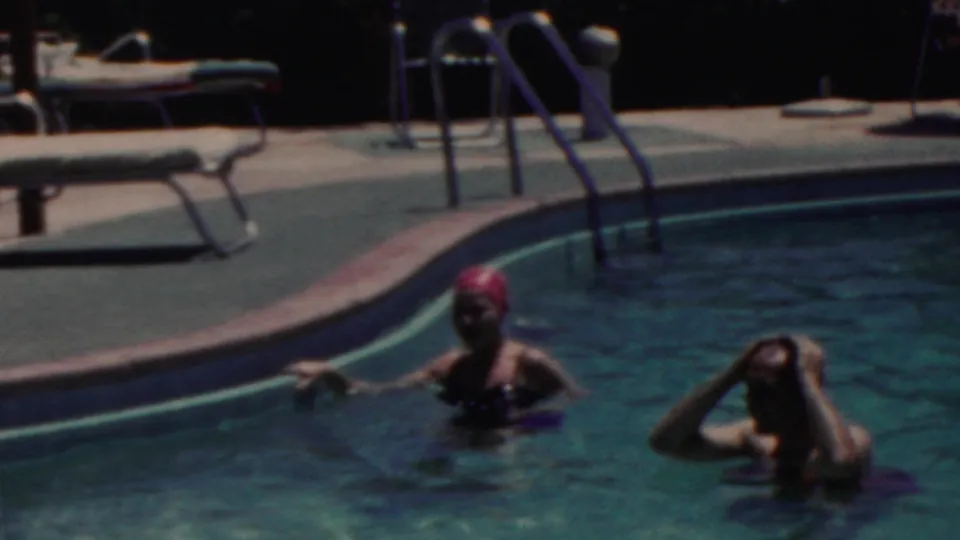 Two adults swim near the edge of a pool in the 1949 Seaside, New Jersey home-movie footage, with a red swim cap, lounge chairs, and a metal ladder visible on the deck.