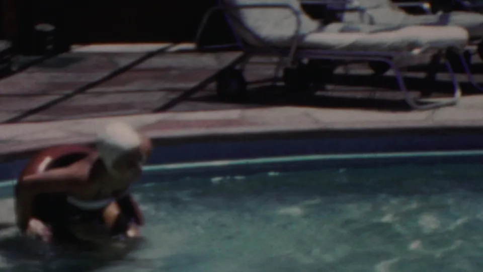 A swimmer in the 1949 Seaside, New Jersey pool footage bends around or holds a small float-like object in the water, with lounge furniture and the pool edge behind them.