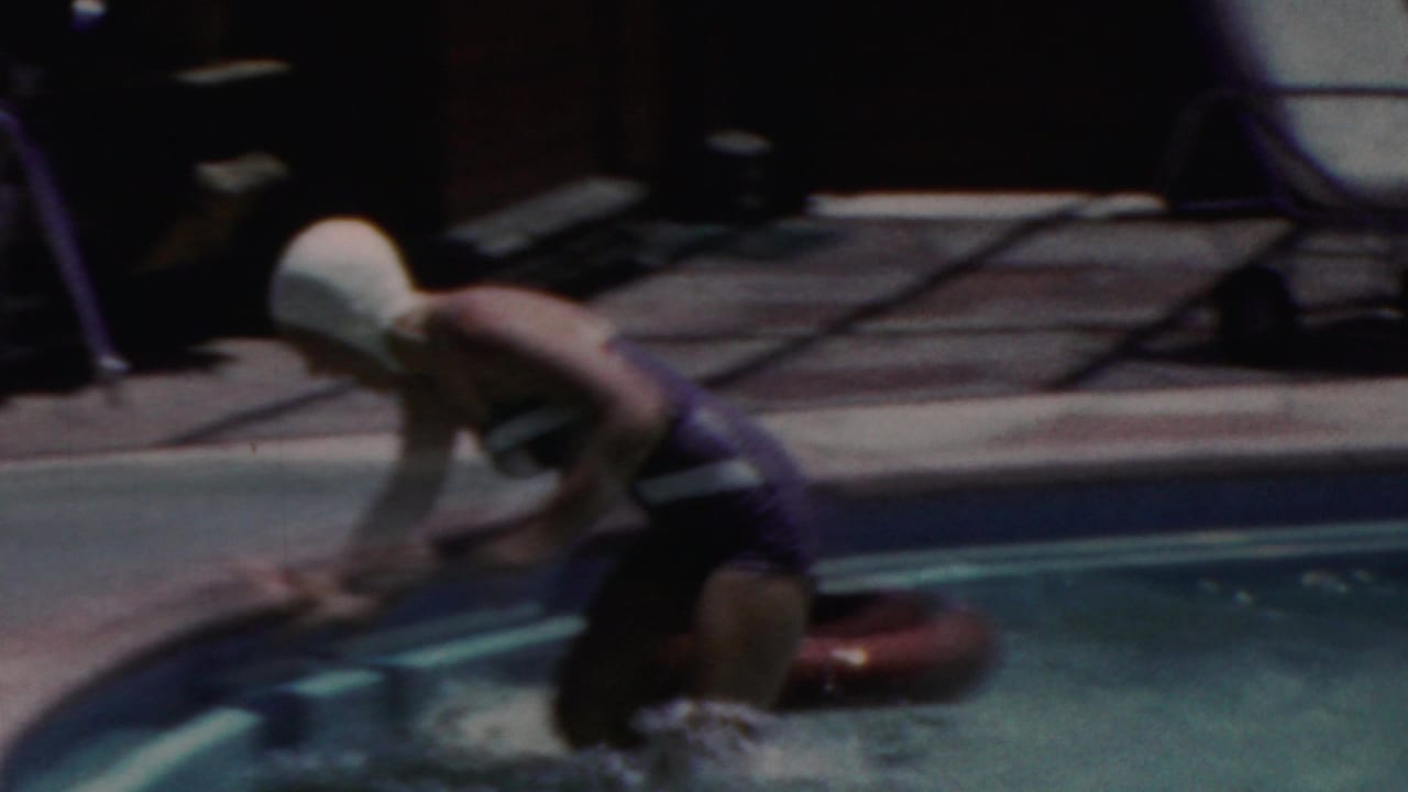 In this 1949 Seaside, New Jersey home-movie frame, a woman in a dark one-piece swimsuit and white swim cap leans into a backyard pool near a red inner tube.