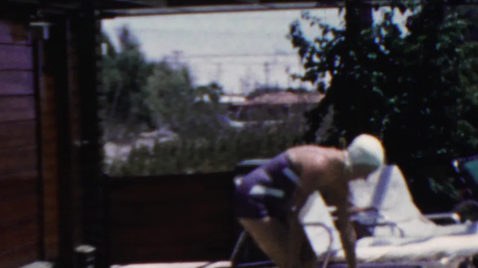 A woman in a white swim cap bends beside a white lounge chair on the pool deck, with trees and nearby buildings in the background of the 1949 Seaside, New Jersey footage.