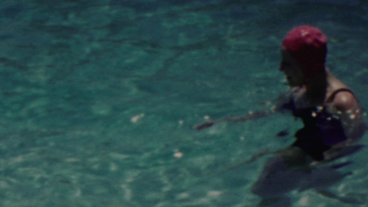 A swimmer in a red cap and dark swimsuit is partly turned in turquoise pool water, with sunlight and film softness obscuring facial detail.