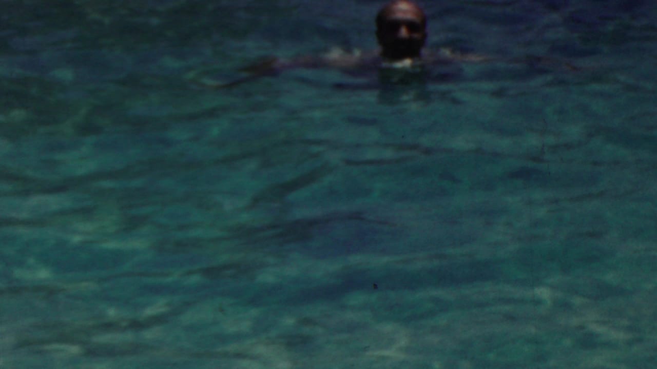 A swimmer is visible from the head and shoulders in a darker section of the pool, with a red-capped swimmer partly visible at the edge of the frame.