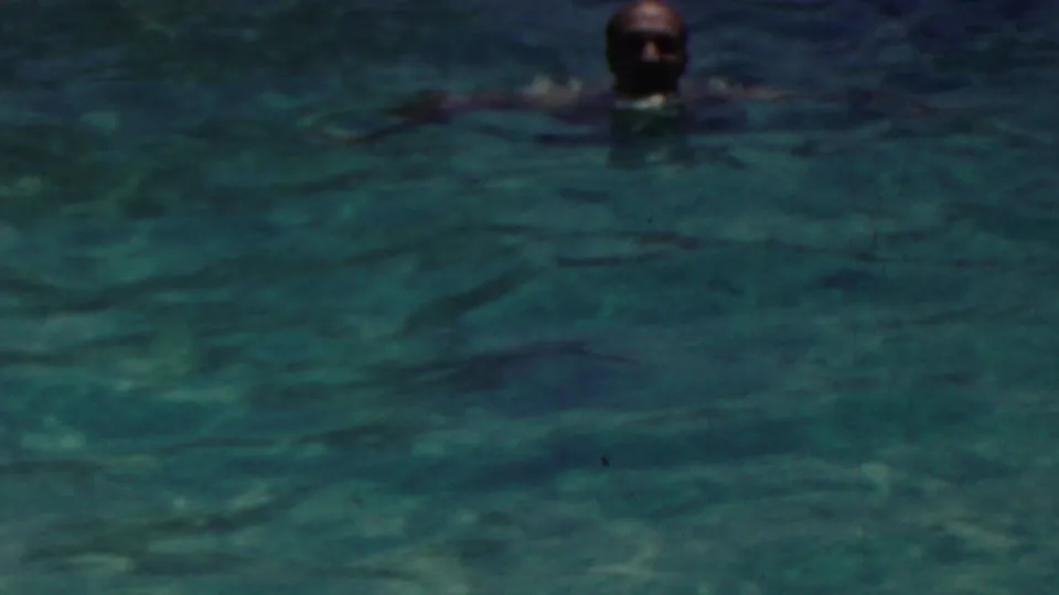 A swimmer is visible from the head and shoulders in a darker section of the pool, with a red-capped swimmer partly visible at the edge of the frame.
