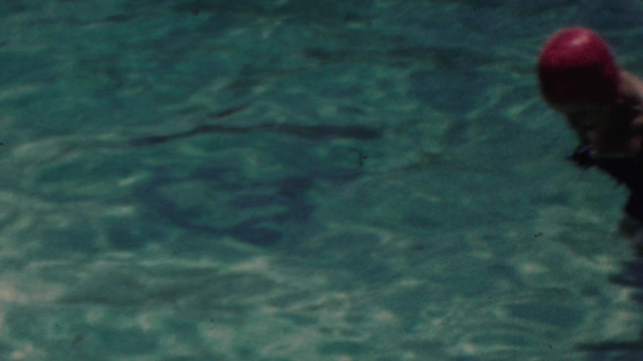 A red-capped swimmer is partly framed at the right edge of turquoise pool water, with bright ripples and soft 8mm color visible across the frame.
