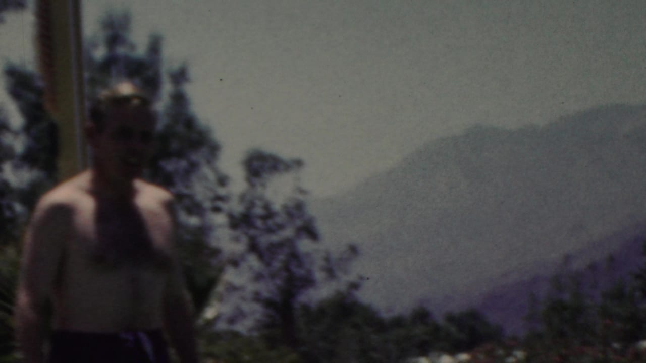 A shirtless man stands near the water in the 1949 Seaside, New Jersey home-movie footage, with dark trees and hazy distant hills or shoreline behind him.