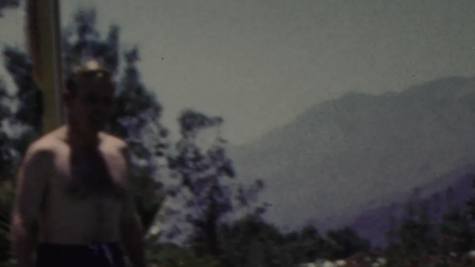 A shirtless man stands near the water in the 1949 Seaside, New Jersey home-movie footage, with dark trees and hazy distant hills or shoreline behind him.