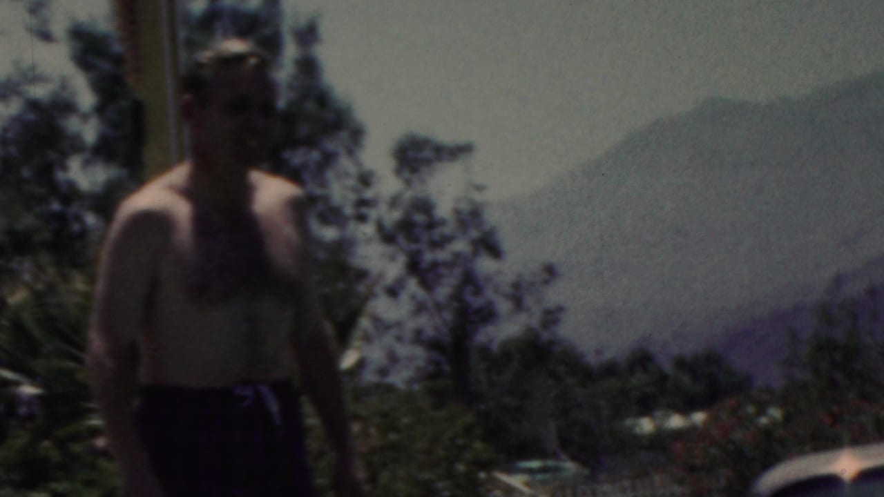 In this 1949 seaside home-movie frame from Seaside, New Jersey, a shirtless adult man in dark swim trunks stands near the water with trees and distant hazy hills in the background.