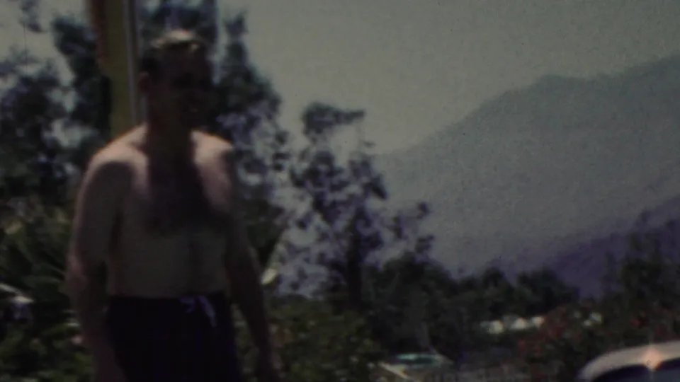 In this 1949 seaside home-movie frame from Seaside, New Jersey, a shirtless adult man in dark swim trunks stands near the water with trees and distant hazy hills in the background.