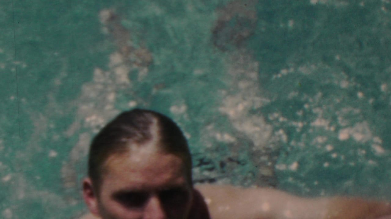 A close, soft-focus home-movie frame shows the swimmer near the camera with rippling turquoise water and splashes behind him.