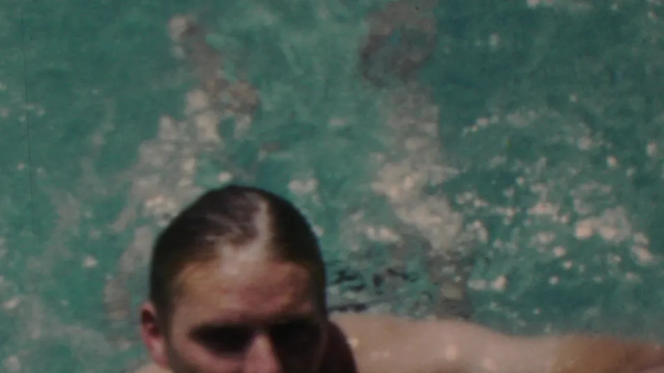 A close, soft-focus home-movie frame shows the swimmer near the camera with rippling turquoise water and splashes behind him.