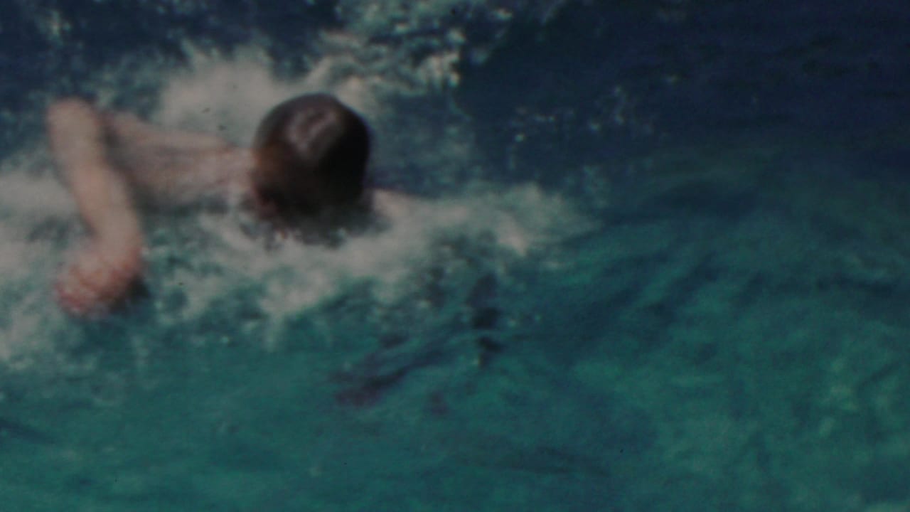 The swimmer is seen from behind as he moves away through darker pool water, with foam and ripples marking his path.