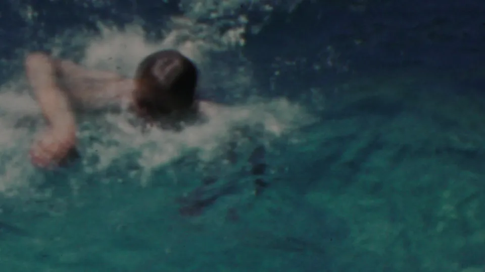The swimmer is seen from behind as he moves away through darker pool water, with foam and ripples marking his path.