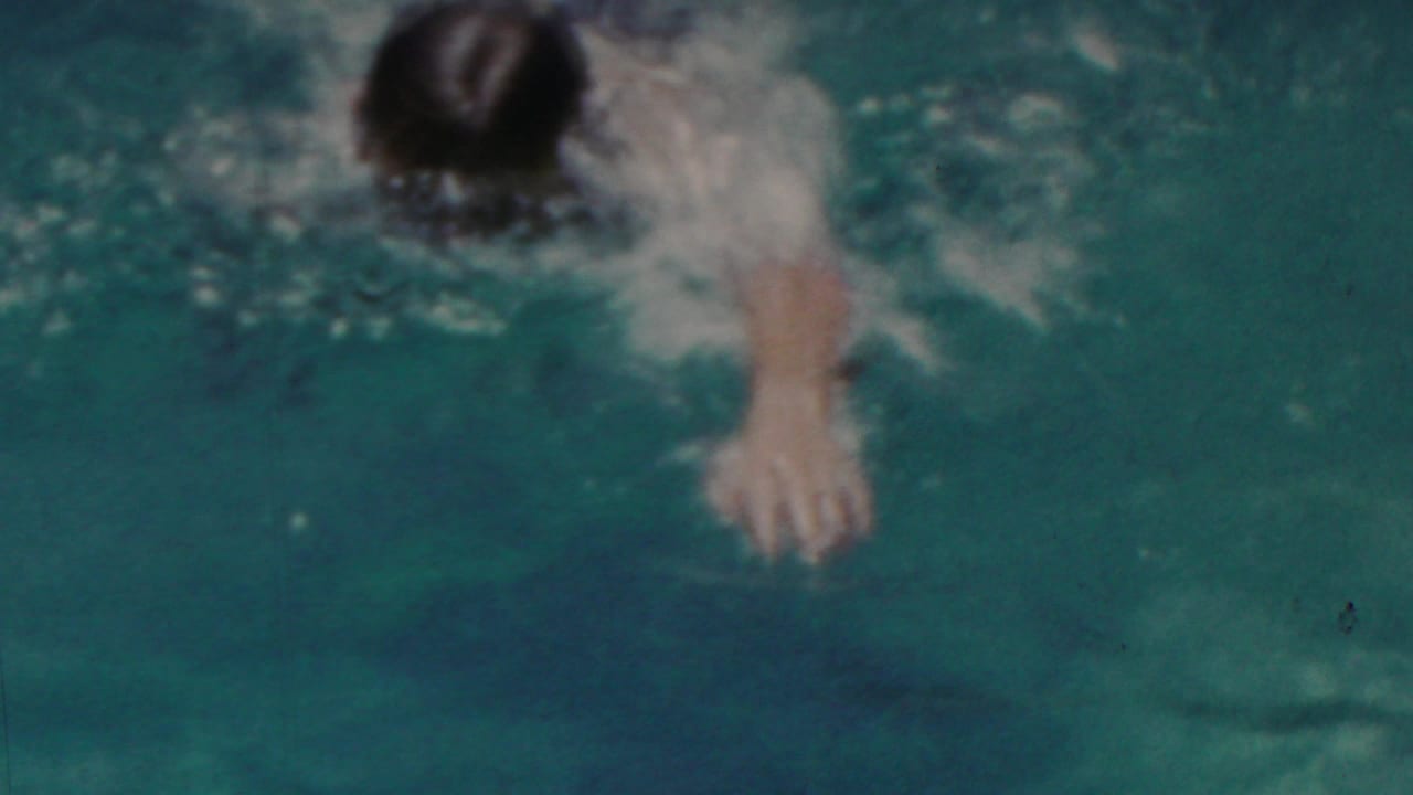 A swimmer moves through blue-green pool water in 1949 Seaside, New Jersey home-movie footage, with splashes and soft 8mm color grain obscuring some detail.