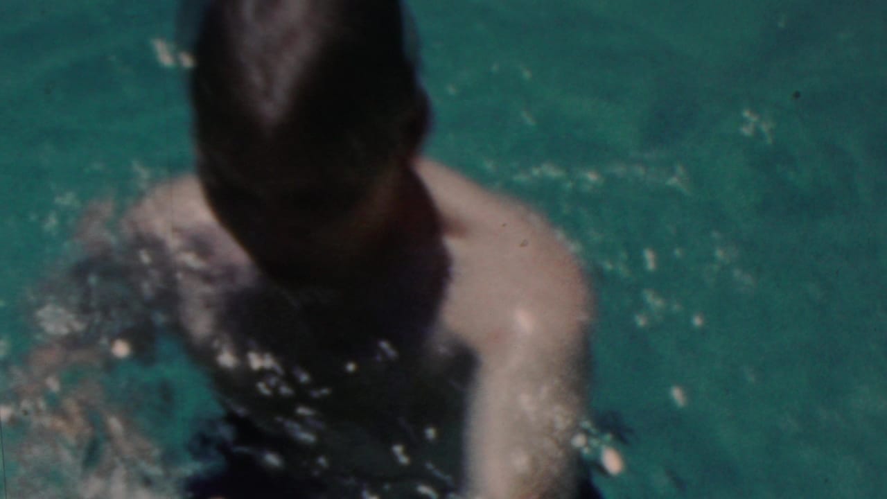 A swimmer with slicked dark hair and bare shoulders is partly submerged in sunlit pool water in the short Seaside, New Jersey home-movie sequence.