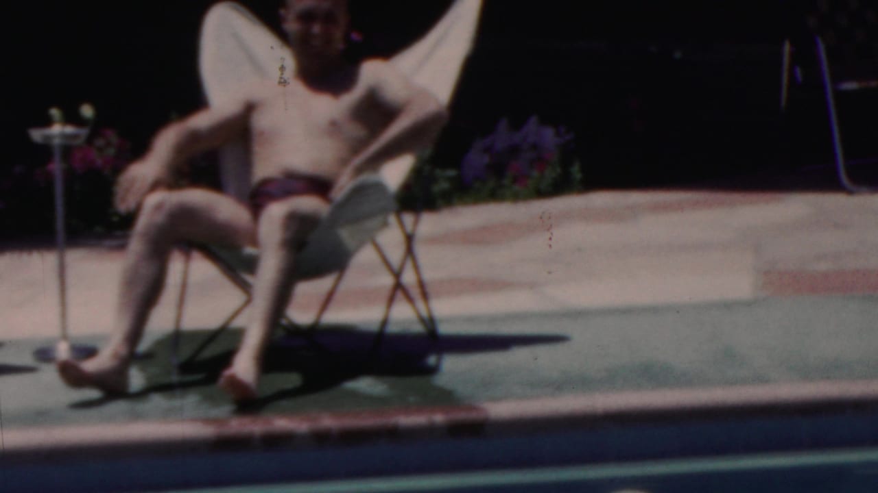 In Seaside, New Jersey, 1949, a shirtless man in dark swim trunks sits in a white folding chair near the edge of a backyard swimming pool.