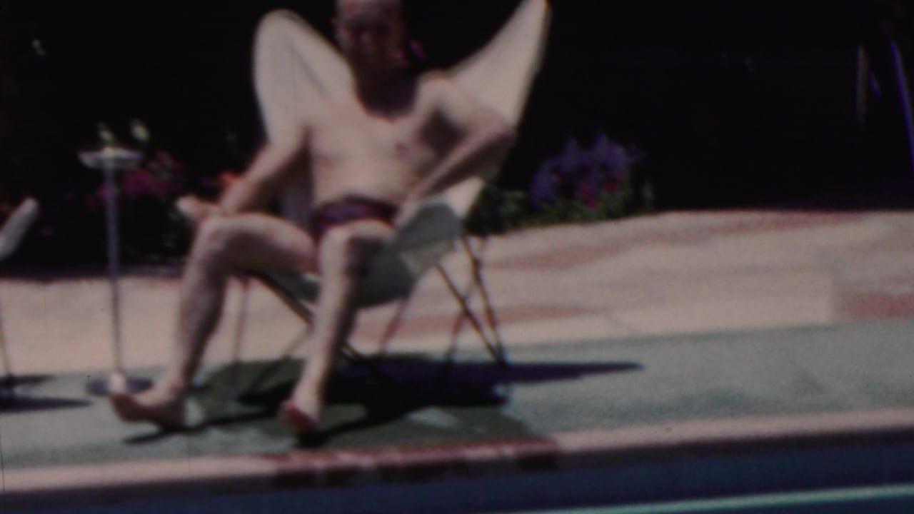 A man sits back in a white folding lawn chair beside the pool, with flowers and a small table visible in the background of this 1949 Seaside home movie frame.