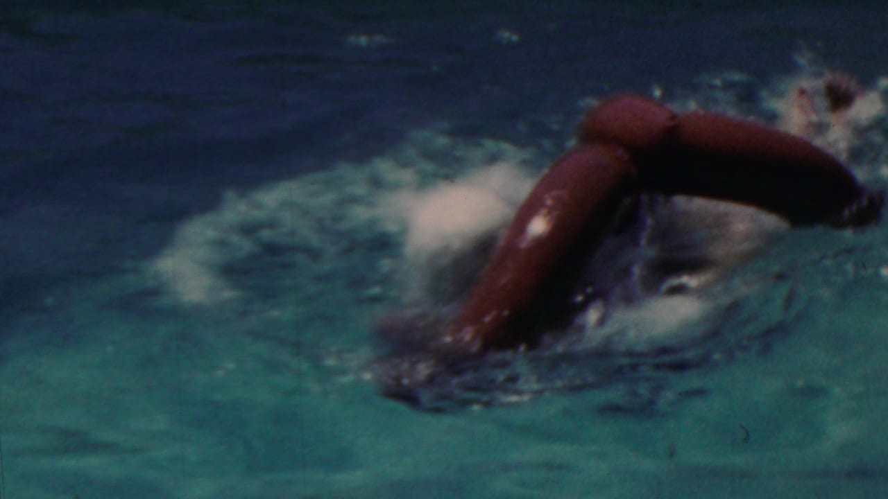 The swimmer turns forward in the pool with the red inflatable ring partly submerged, leaving splashing water and motion blur across the 8mm frame.