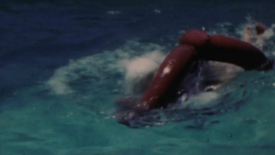 The swimmer turns forward in the pool with the red inflatable ring partly submerged, leaving splashing water and motion blur across the 8mm frame.