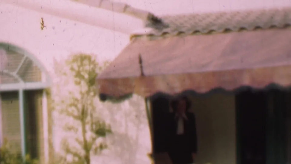 In this 1955 San Diego home-movie frame, a woman in a dark outfit and white blouse is partly visible beneath a scalloped awning at a residential doorway.