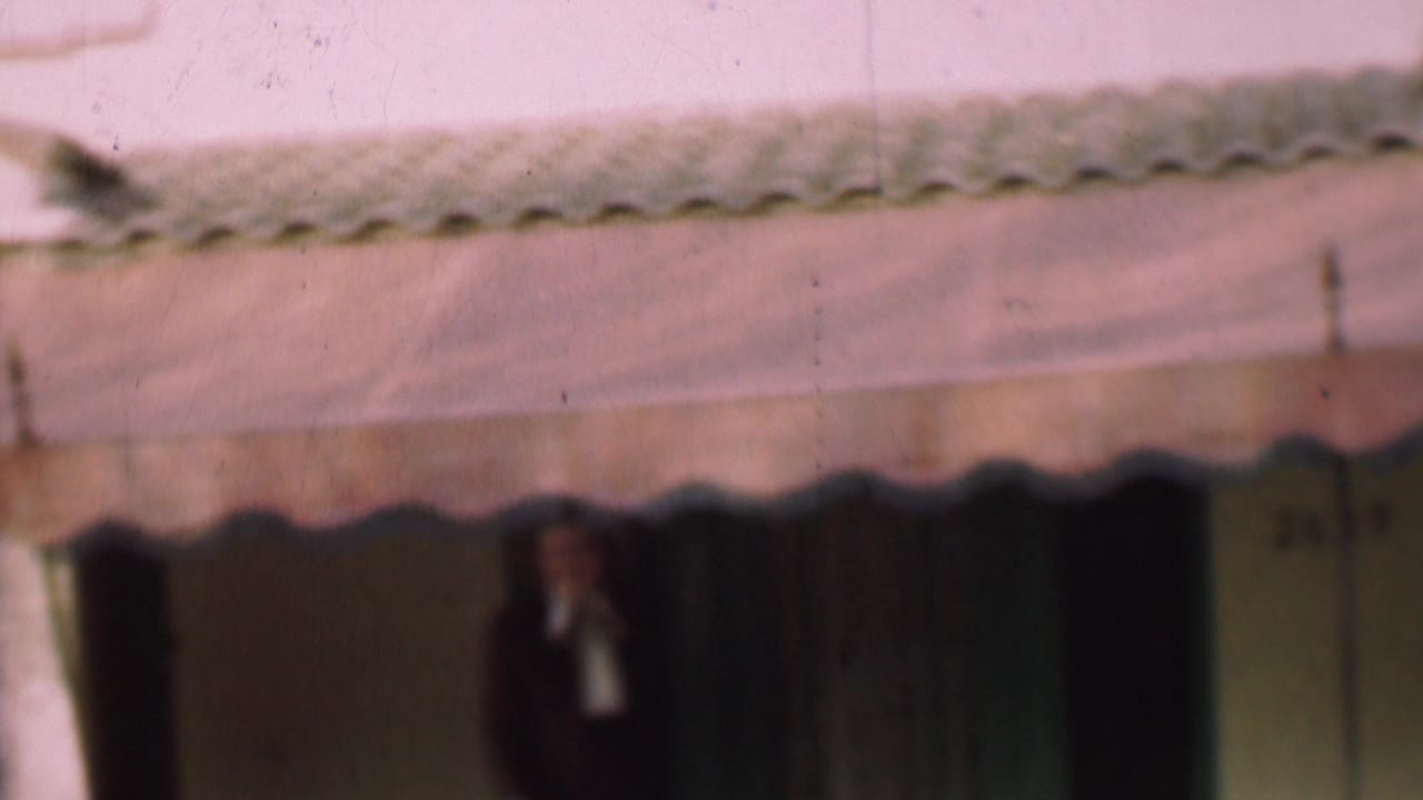 The woman remains under the awning outside the San Diego house, with a dark doorway, curtain area, and green-toned trim visible around her.