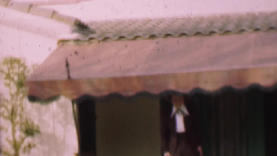 A woman in a dark outfit and white blouse steps out from under the scalloped awning of a residential exterior in the 1955 San Diego home-movie footage.