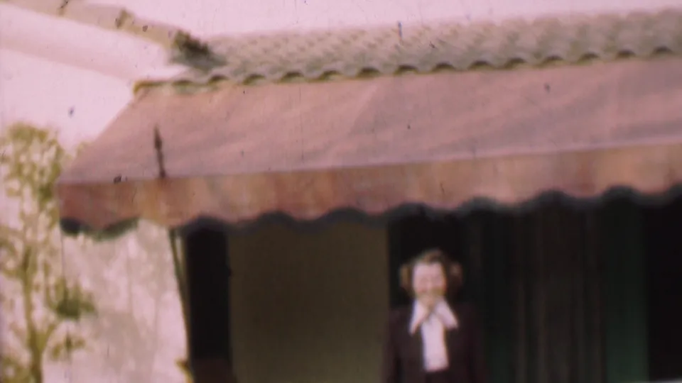 The woman appears to smile toward the camera near the doorway, with the scalloped awning, wall surface, curtains, and green-toned trim still visible in the 1955 San Diego scene.