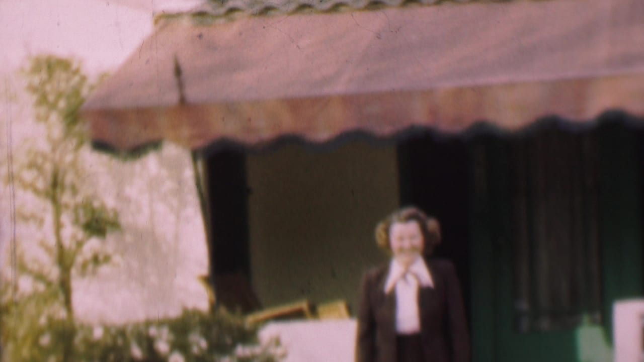In supplied 1955 San Diego home-movie footage, a smiling adult woman in a dark suit and white bow blouse stands near a doorway beneath a scalloped awning.