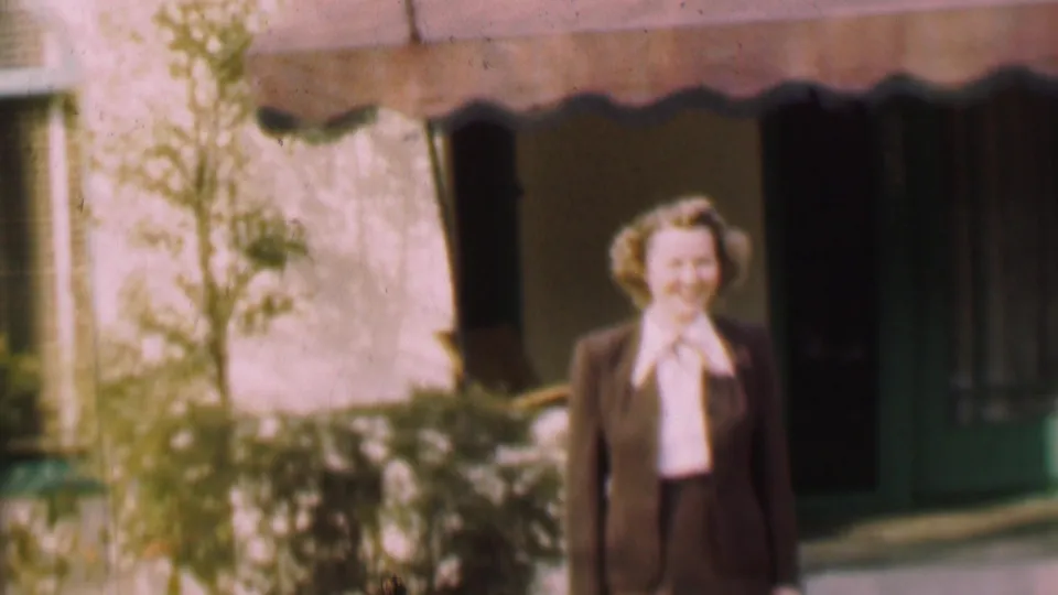 The woman walks closer in the 1955 San Diego exterior scene, with her dark suit, white bow blouse, scalloped awning, doorway, and surrounding shrubs visible.