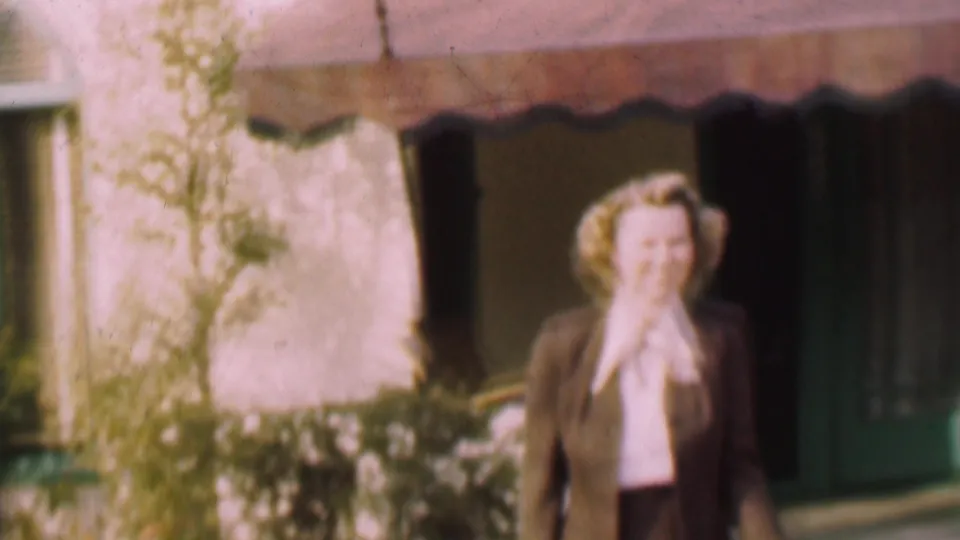 A closer soft-focus frame shows the smiling woman lifting a hand near her face while standing by the awning, doorway, pale wall, and garden plantings.
