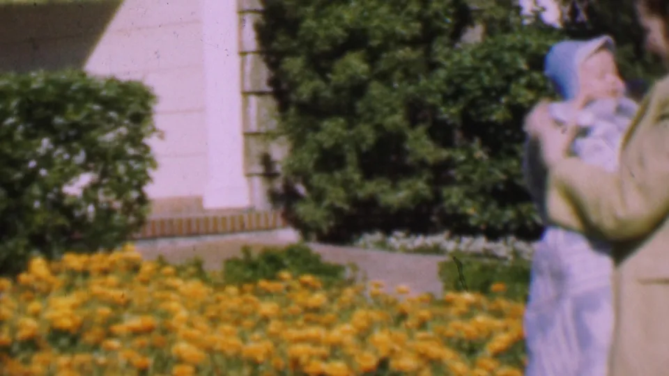 1955 San Diego 8mm frame showing a woman near the house exterior holding a bundled baby beside yellow flowers and shrubs.