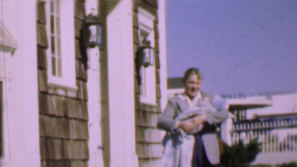 1955 San Diego home-movie frame of a woman on a residential porch holding a bundled baby, with house siding, windows, and blue sky visible.