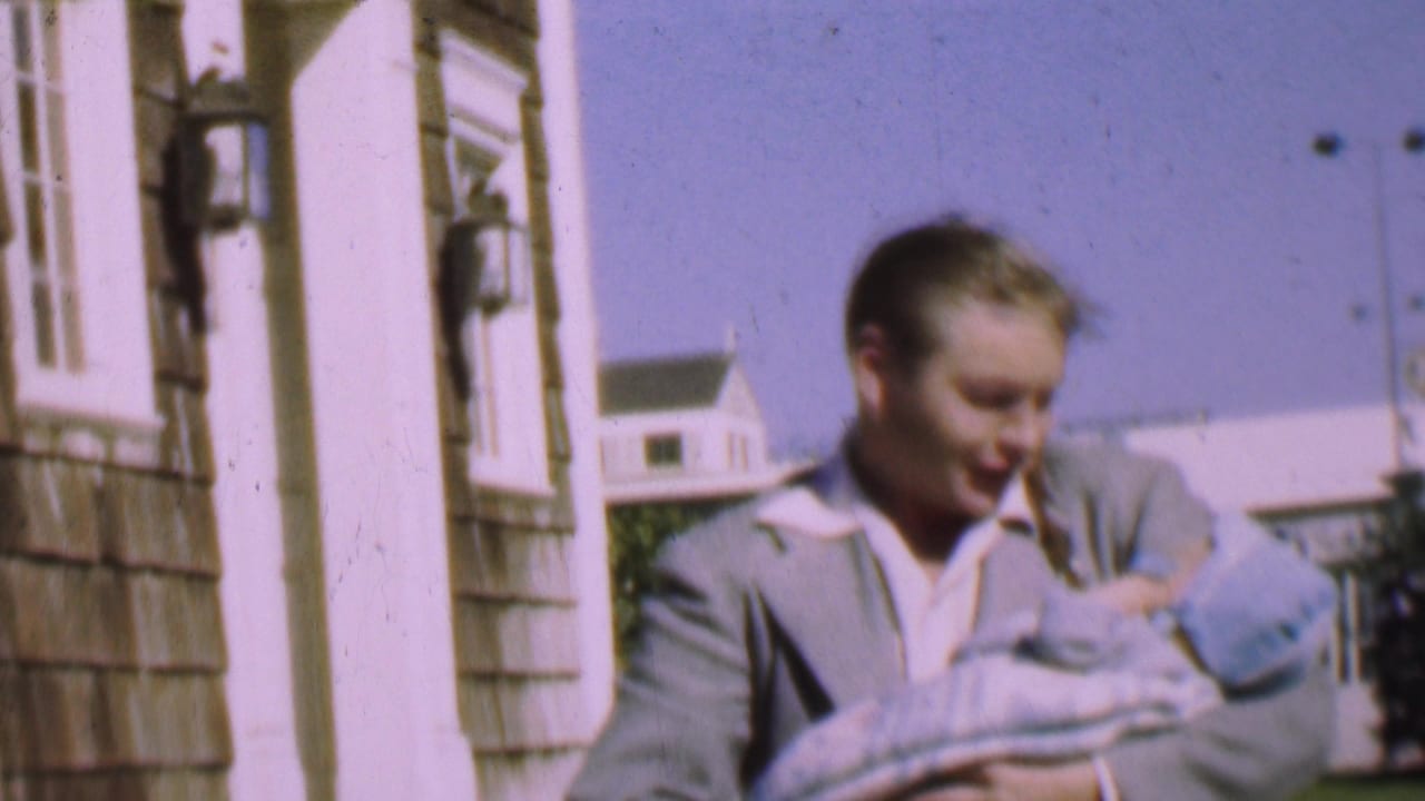In 1955 San Diego home-movie footage, a young man in a light jacket holds a bundled baby near shingled siding and white exterior trim.