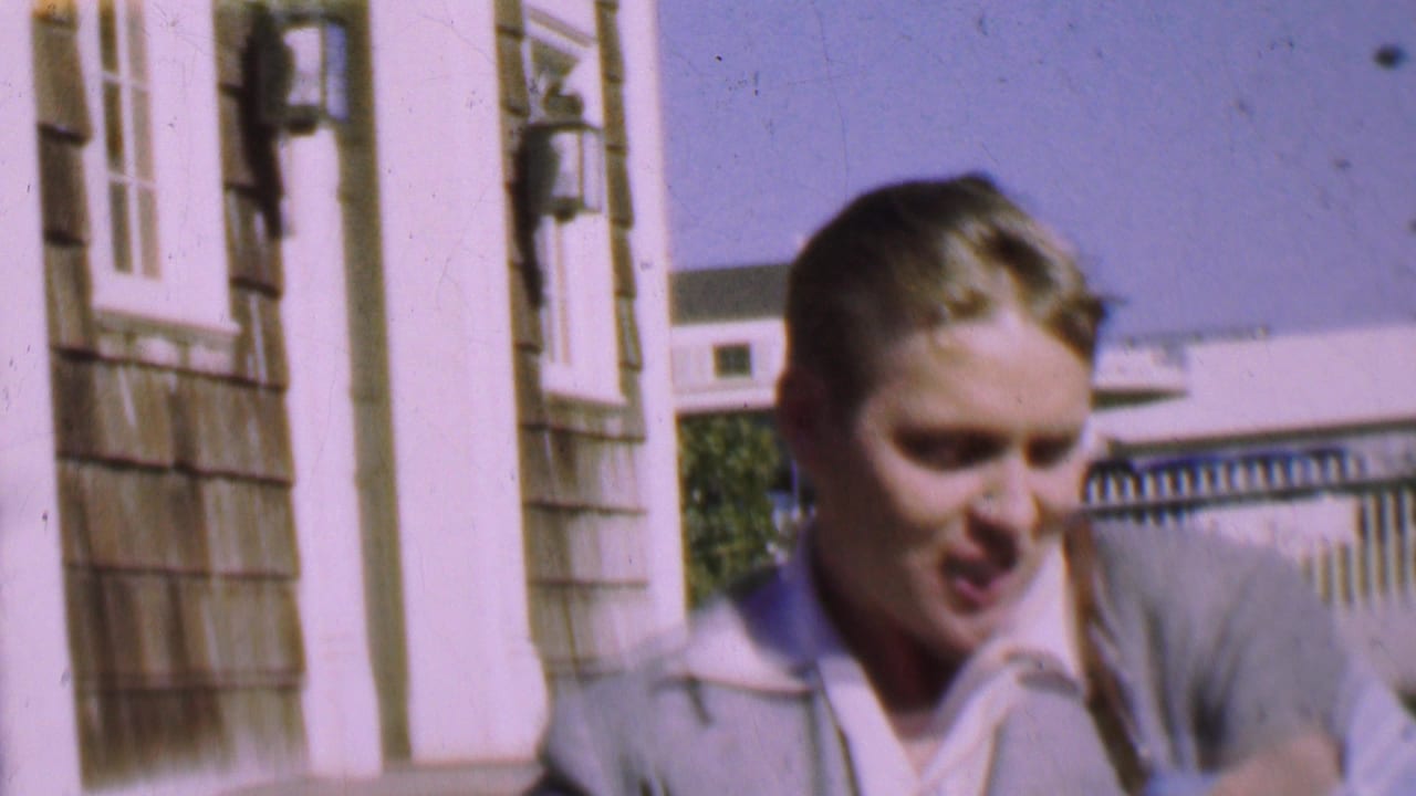 The young man smiles close to the camera outside a residential-looking exterior, with white trim, shingled siding, and a pale building in the background.