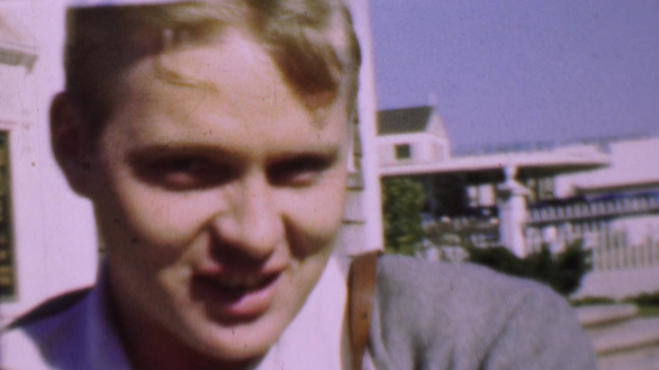 Close view of a young man's face with a house and railing behind him.