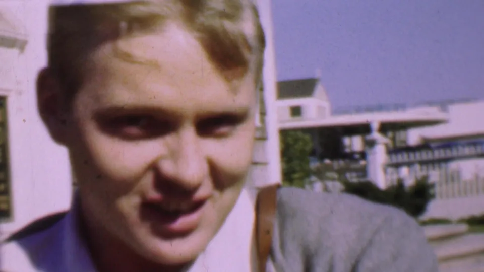 A close, softly focused view shows the young man facing the camera, with a residential exterior, railing, and pale building visible behind him.