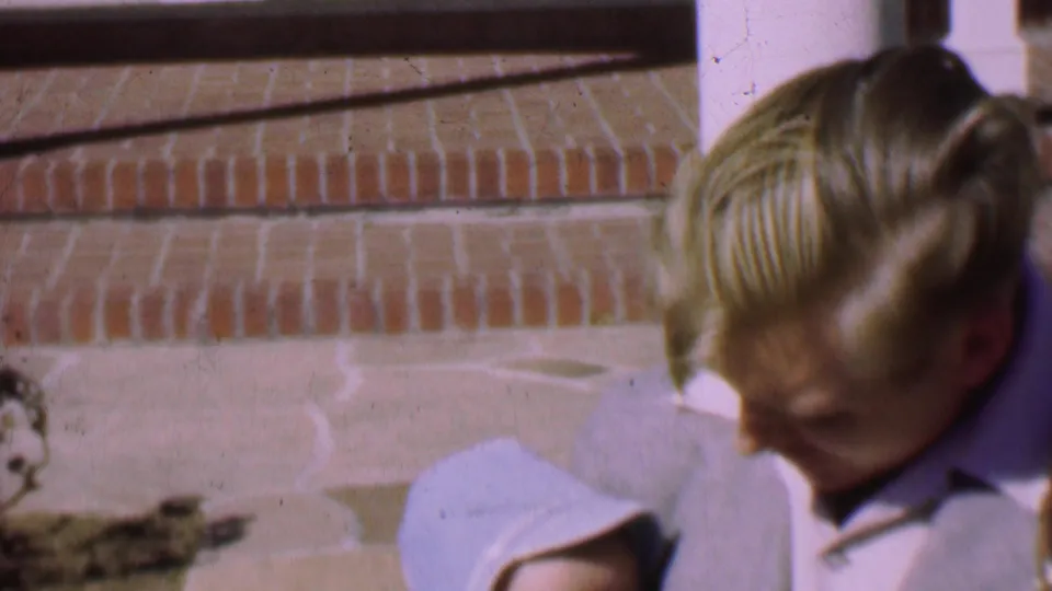 The young man looks down toward the bundled baby beside brick steps and a white-trimmed house exterior in the 1955 San Diego home-movie scene.