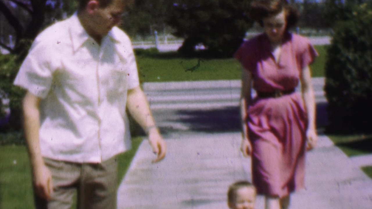 1955 San Diego home-movie frame showing a man in a white short-sleeved shirt and a woman in a pink dress walking on a residential sidewalk with a small child partly visible near the bottom edge.