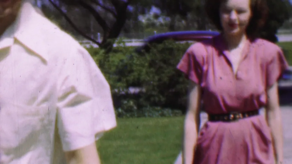 The woman in a pink dress and dark belt faces the camera on the sunny sidewalk, with the man in a white shirt partly cropped at the left edge and lawns and shrubs behind her.