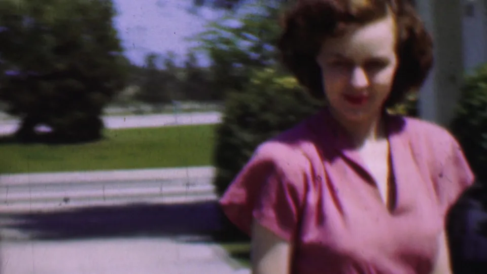 Close 1955 San Diego home-movie frame of the woman in a pink dress near the right side of the sidewalk, with soft focus, lawn, shrubs, and a pale background line behind her.