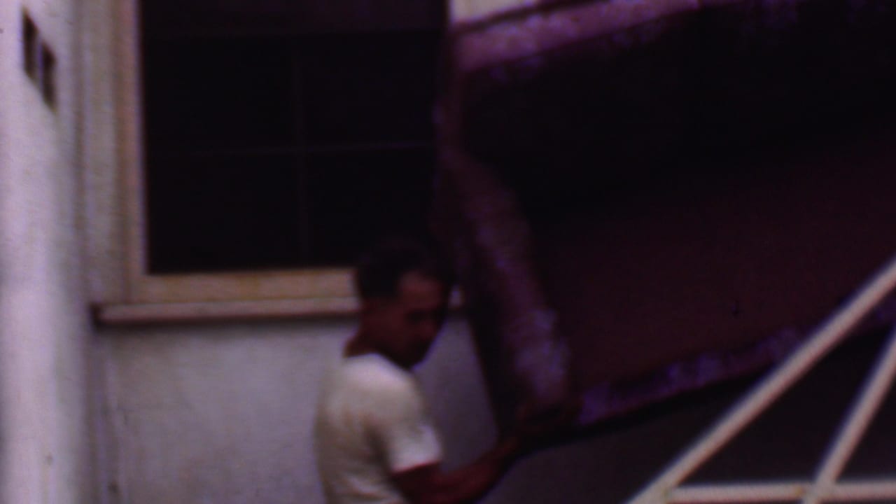 In 1956 Phoenix home-movie footage, a man in a white T-shirt maneuvers a large upholstered couch beside exterior stairs and a stucco wall.