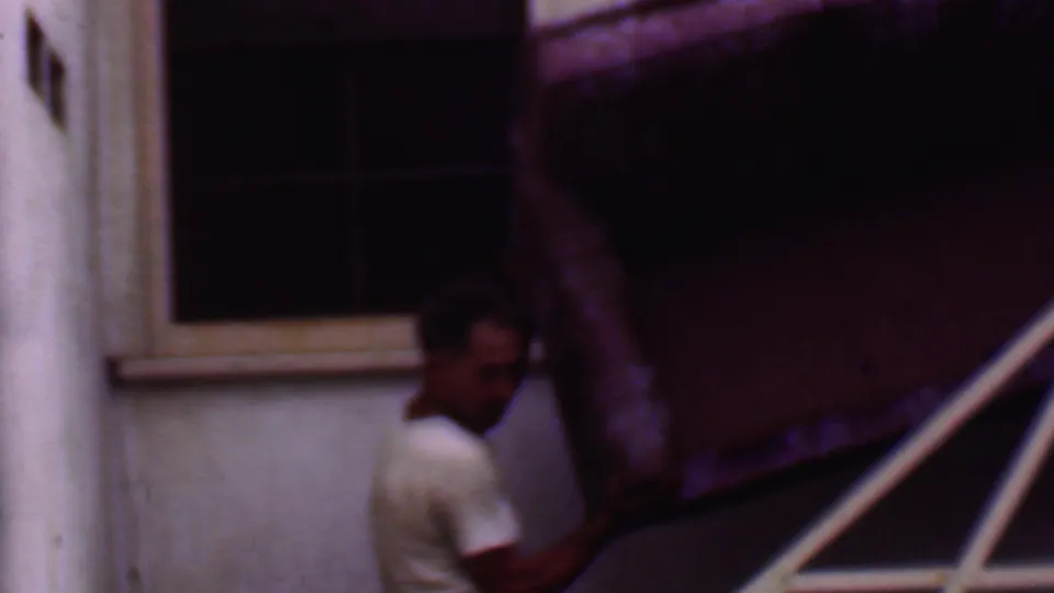 In 1956 Phoenix home-movie footage, a man in a white T-shirt maneuvers a large upholstered couch beside exterior stairs and a stucco wall.