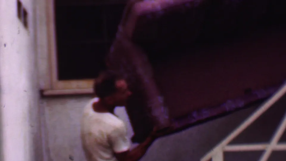 A man in a white T-shirt is partly obscured by a large couch as he carries it along outdoor stairs in the documented Phoenix 1956 setting.
