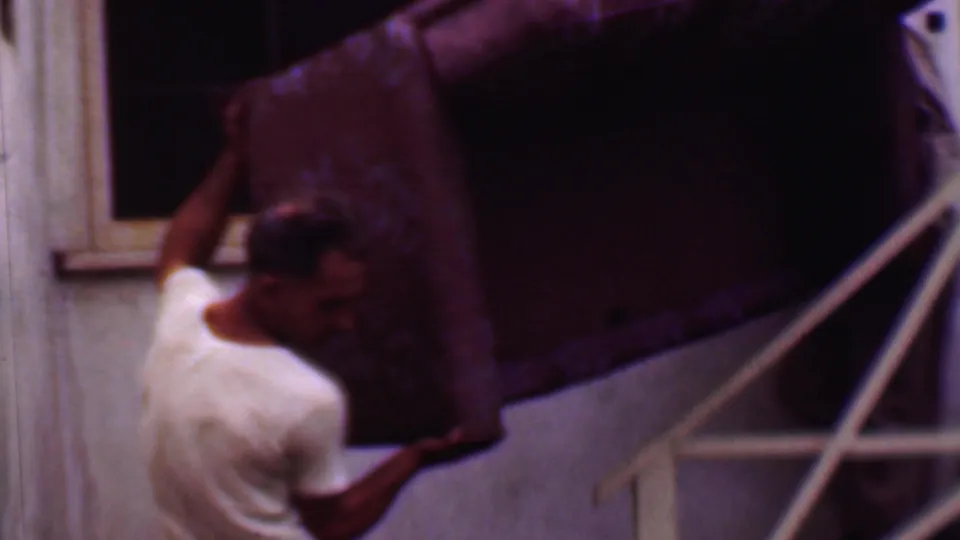 The home-movie frame shows a man straining with a large upholstered couch on exterior stairs beside a stucco building wall and window.
