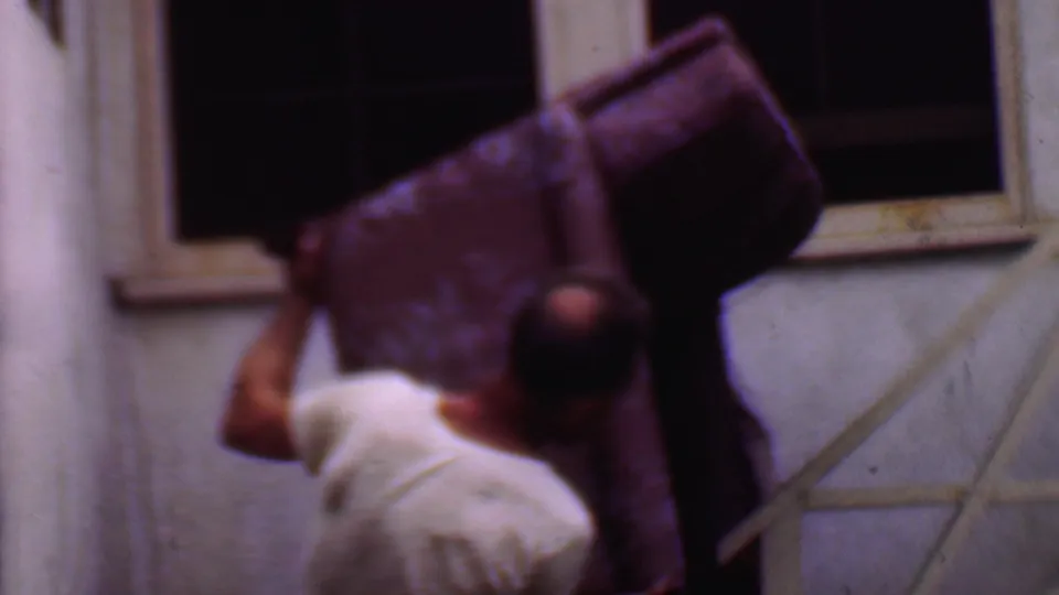 A rear view shows the man guiding the large couch down exterior stairs, with a window, railing, and stucco wall visible in the 1956 Phoenix home-movie scene.