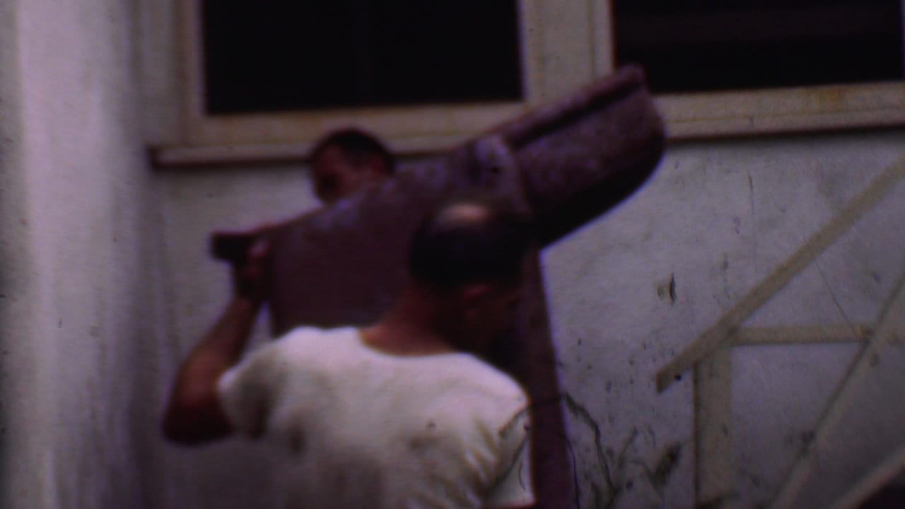 In Phoenix, Arizona, 1956 home-movie footage, two adult men carry a large purple patterned couch near a narrow outdoor stairway and stucco wall.