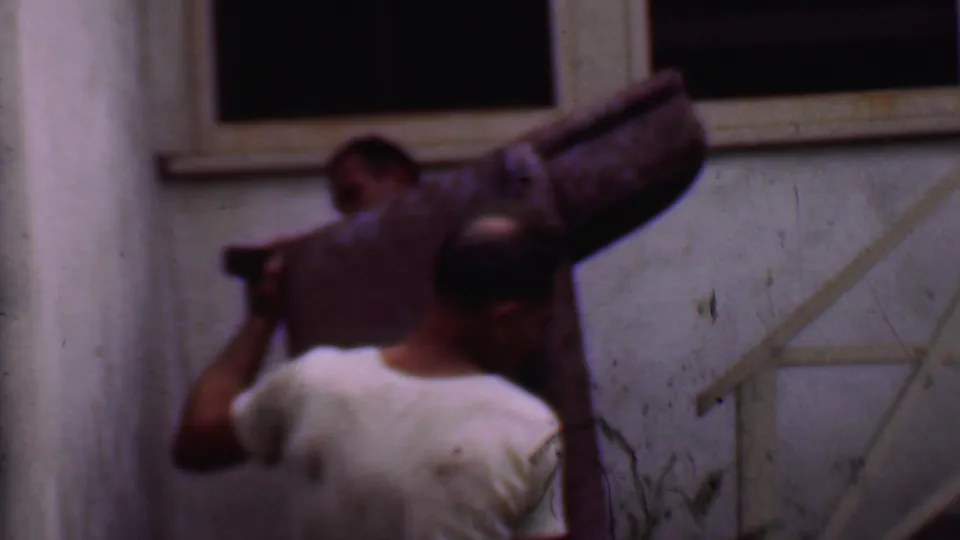 In Phoenix, Arizona, 1956 home-movie footage, two adult men carry a large purple patterned couch near a narrow outdoor stairway and stucco wall.